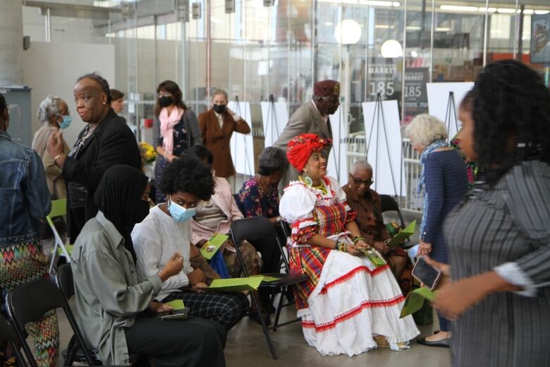 A scene of a crowd of people at the Hamilton Farmers Market, chatting and discussing Dr. Ethilda Tillie Johnson's work and history at the market. Half of the attendees are sitting, the others are standing and holding pamphlets of Johnson's brief history.