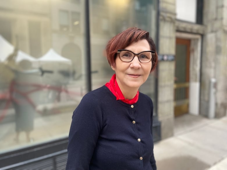 A middle-aged woman with glasses stands on a city sidewalk and smiles.