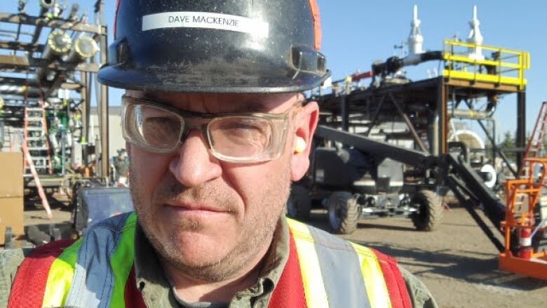 A man with a hard hat and safety vest stands in a yard with a forklift behind him. 