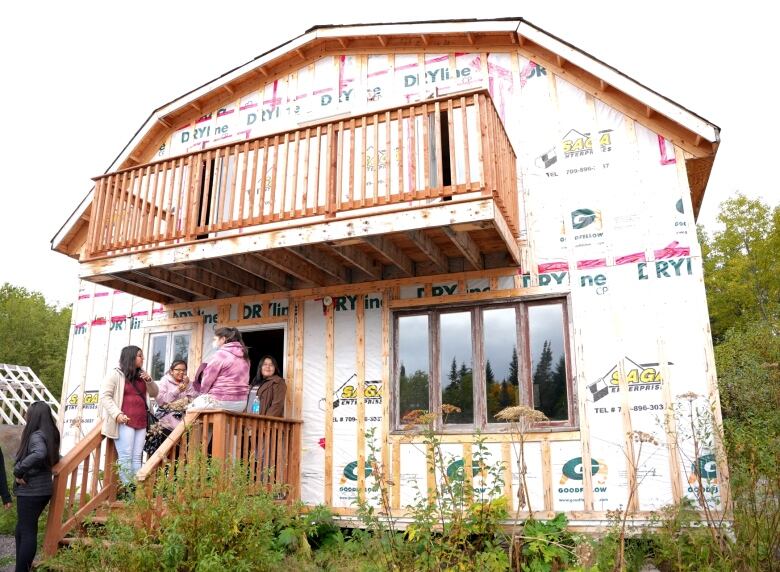 A two story cabin is covered in white builders paper with four students standing in its entry way. 