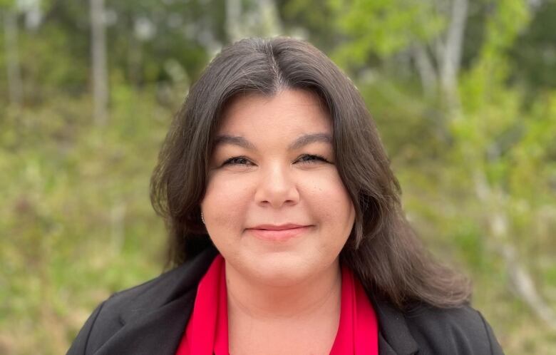 Stacy Wight looks straight into the camera, wearing a red blouse, with trees in behind her. 