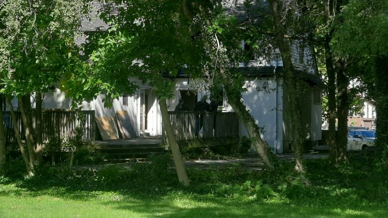 A white house is seen from a distance, surrounded by trees. Two people stand on the porch. 