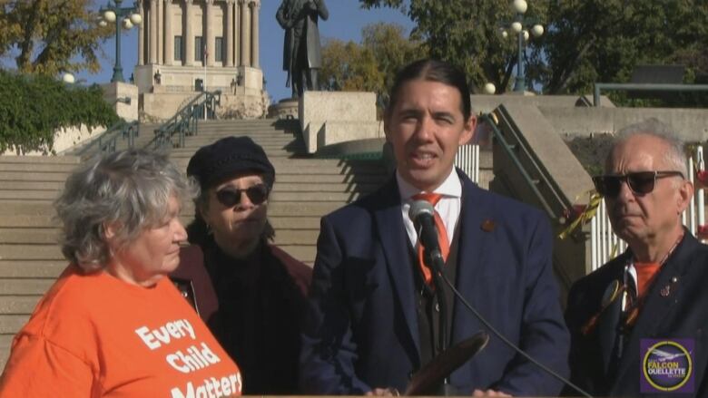 A man stands speaking into a microphone on a staircase with people around him. In the background, the Manitoba Legislative Building can be seen.