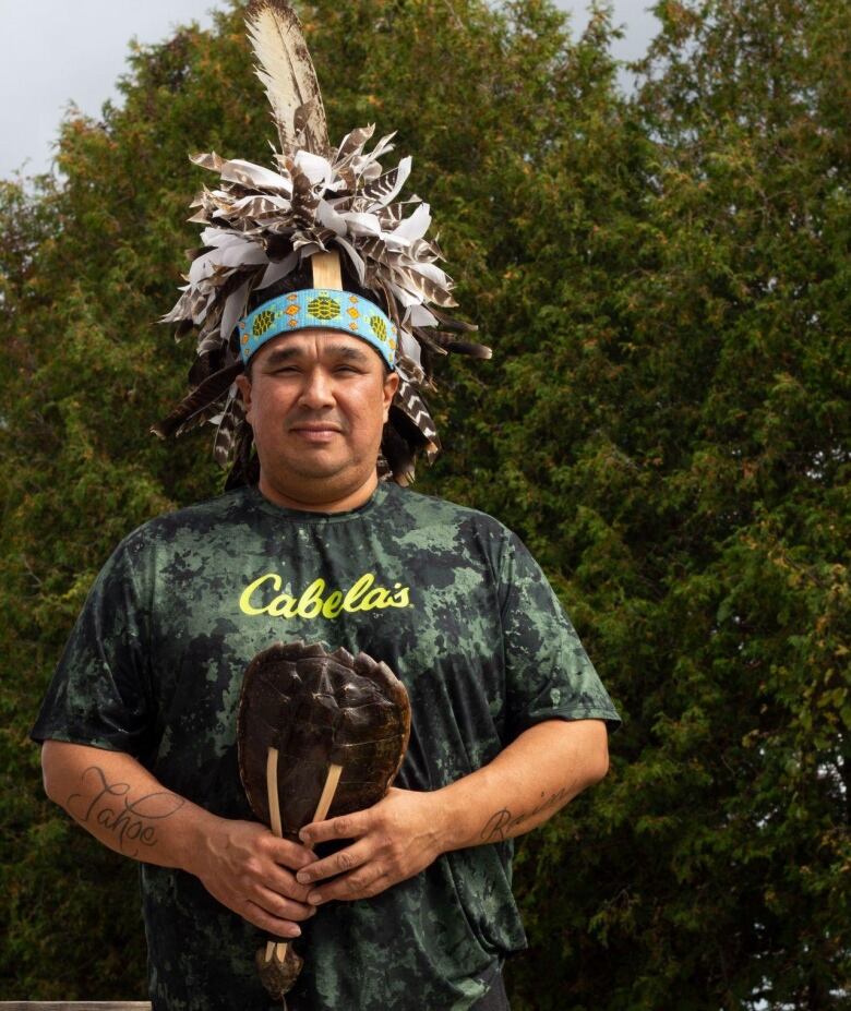 A man wears an elaborate feather headdress and holds a turtle shell in his hands.