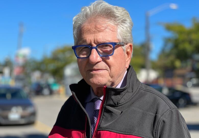 Man with white hair and glasses and a dark coat faces the camera.