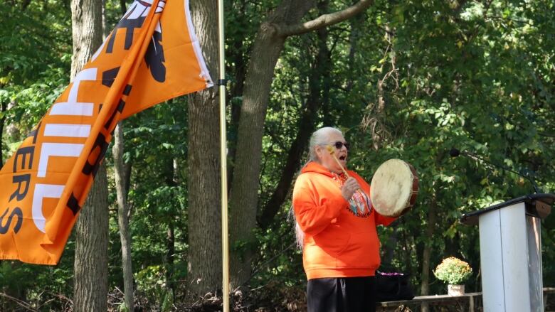 Woman stands beside a flag, holding a handheld drum and drumstick. 