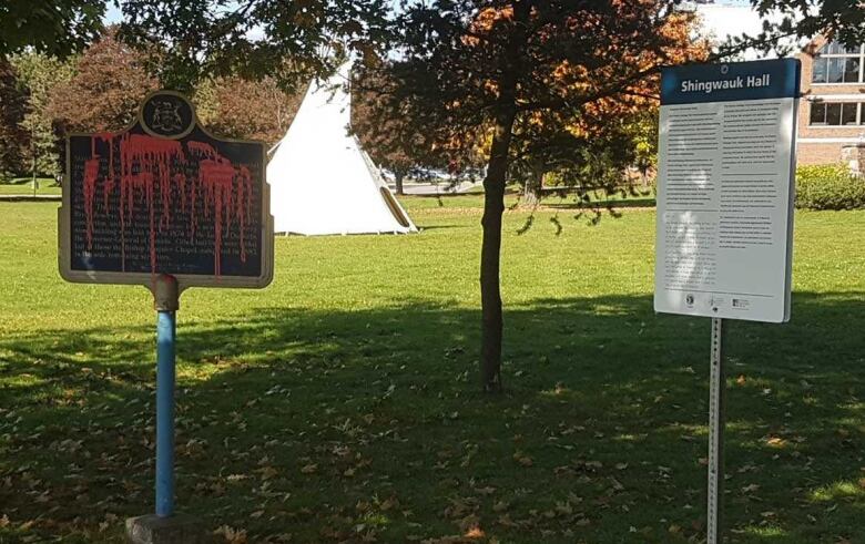 Historic plaque splattered with red paint alongside a white sign with a teepee in the background.