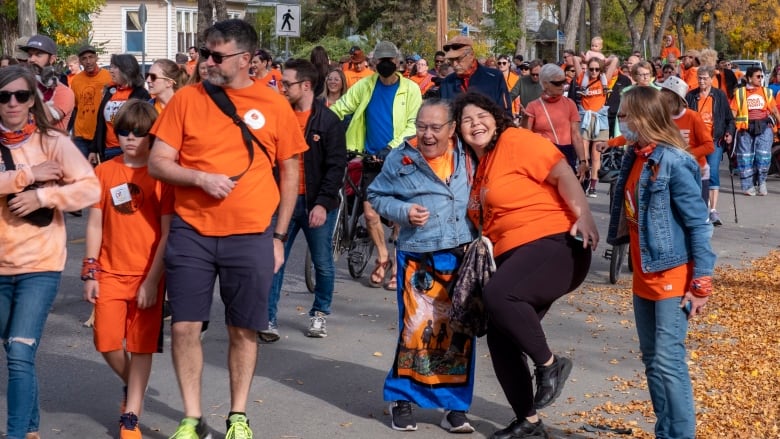 People wearing orange shirts walk on a street, stepping over fallen yellow leaves. 