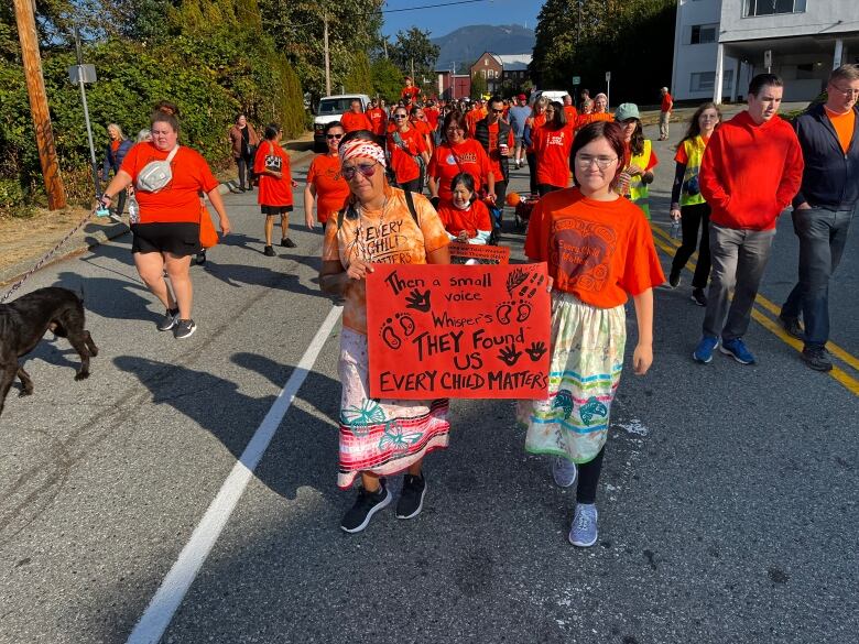 Groups of people marching wearing orange garments. Someone at the front of the line has a sign that reads 'Then a small voice whispers They Found Us Every Child Matters'.