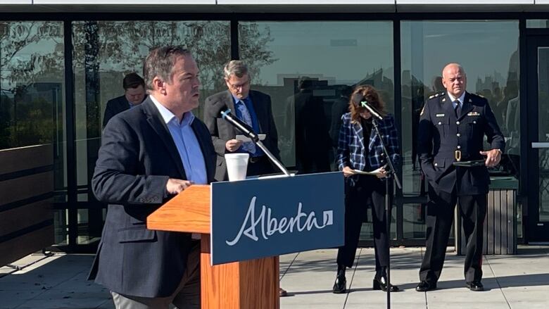 A man is shown speaking into a microphone that's set up on a podium. A mostly filled Styrofoam cup sits on the podium. There are several people standing in the background reading a report. Edmonton's chief of police is also standing in the background, holding his cap at his side with his left hand.