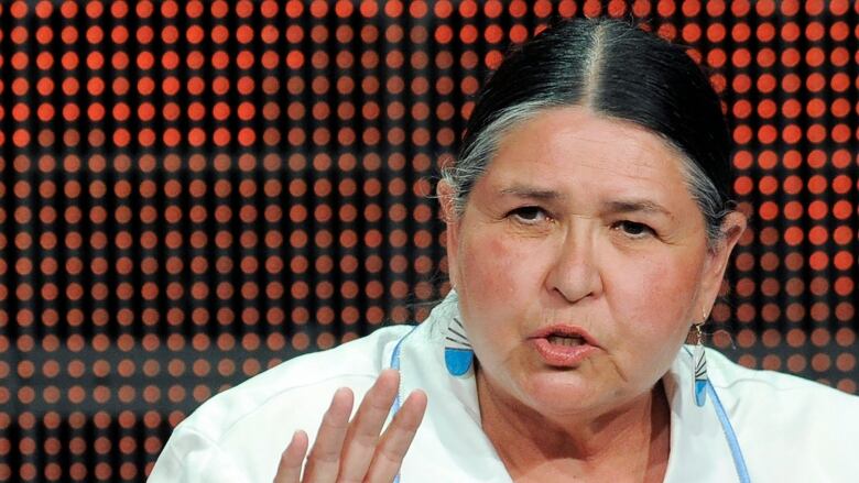 An older woman with blue feather earrings and a white jacket with yellow embroidery speaks towards the camera. 