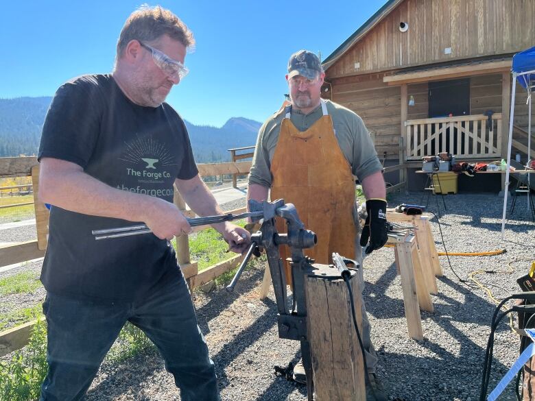 A man holds metal instruments over a small stump, as another man looks on.