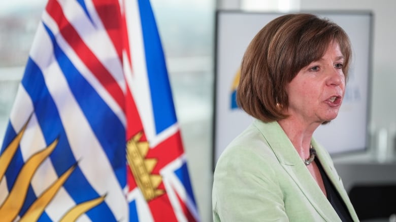 A woman in a green suit with a B.C. flag behind her speaks to reporters who are off camera.