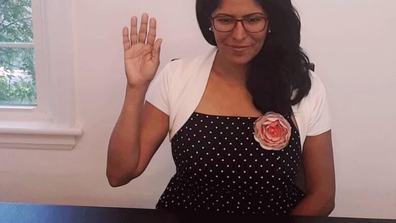 A woman in a white dress smiles in front of a computer and raises her right hand. 