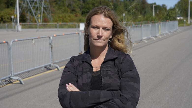 A woman crosses her arms and looks into the camera with cement lot and metal gates in background.