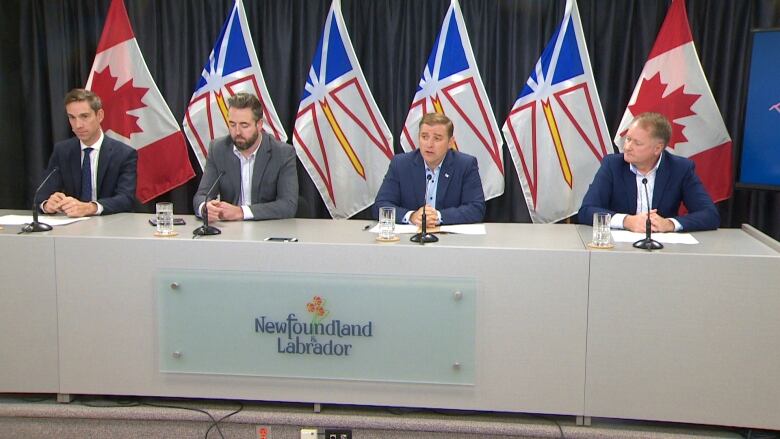 Four men sit at a table in front of microphones for a press conference. There are six flags standing behind them, two Canada flags and four Newfoundland and Labrador flags.