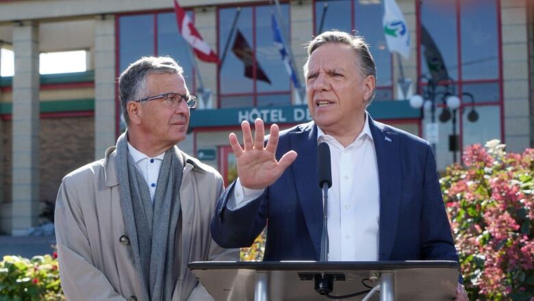 Two man outside. One of them is standing behind a podium and talking.