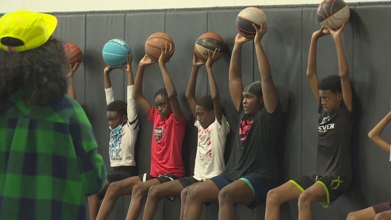 Row of kids seated against a wall holding basketballs above their heads. One of several drills during basketball training at Ballers Union Training Centre in North York.