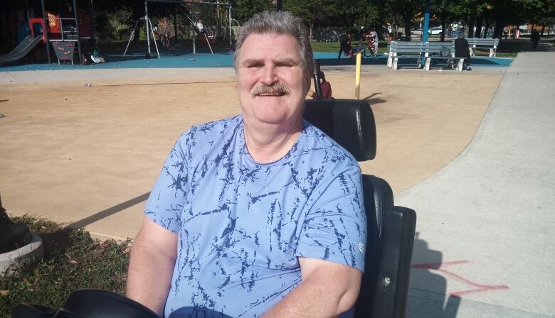 A man with short, white hair smiles at the camera. He is sitting in an electric wheelchair in a park.