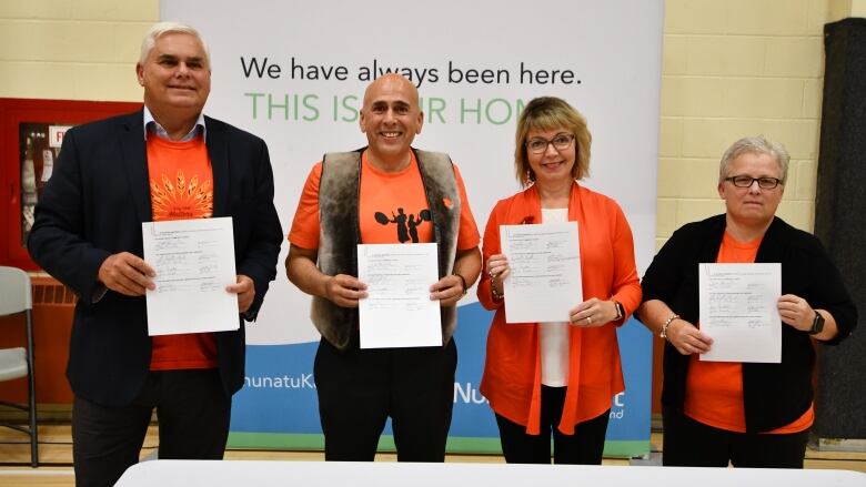 Four people in orange shirts hold up pieces of paper in front of a NunatuKavut banner. 