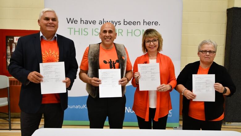 Four people in orange shirts hold up pieces of paper in front of a NunatuKavut banner. 