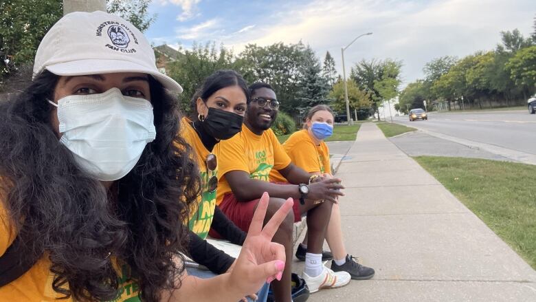 Four people in yellow shirts are sitting on some sort of ledge beside a sidewalk, smiling at the camera, in what looks like a residential area.