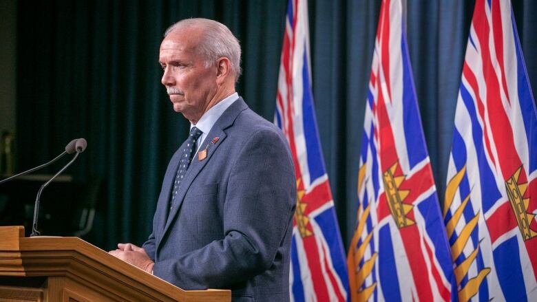 B.C. Premier John Horgan, a white man with a white moustache, looks pensively in front of a few flags of British Columbia.