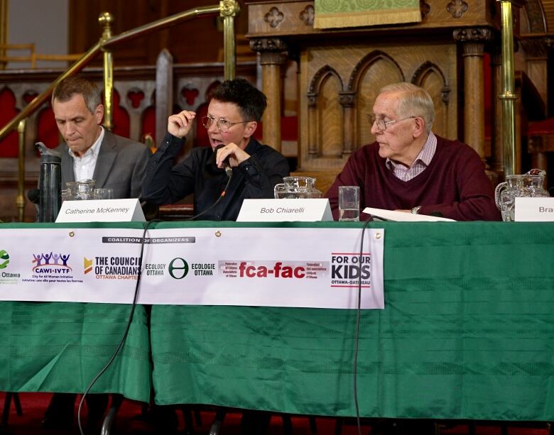 Three politicians sit at a table. The middle one is speaking on a microphone.