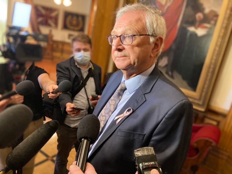 A portrait of a man in the foreground with five microphones in front of him and reporters in the background.