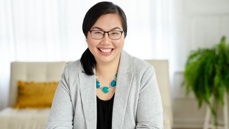 A woman with dark hair in a low ponytail is smiling. She has on a blazer and a necklace and glasses.