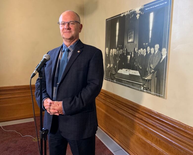 A person stands, with his hands folded, in front of a microphone. On the wall behind him hangs a black and white photo depicting a group of people standing around a table.