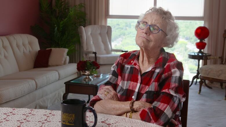 An elderly woman sits in a living room with her arms crossed.