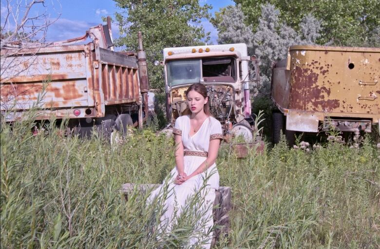 A woman in a white dress sits in a grassy field, with rusted trucks in the background.