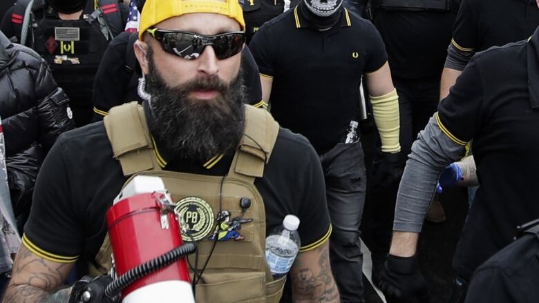 A man wearing sunglasses and a beard with a yellow cap, holding a bullhorn. 