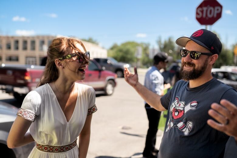 A smiling bearded man in a baseball cap stands outside beside a laughing woman in a white dress and sunglasses.