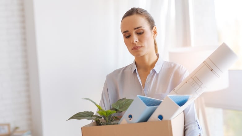 A woman with her hair pulled back looks down as she carries a box of office materials and a plant.