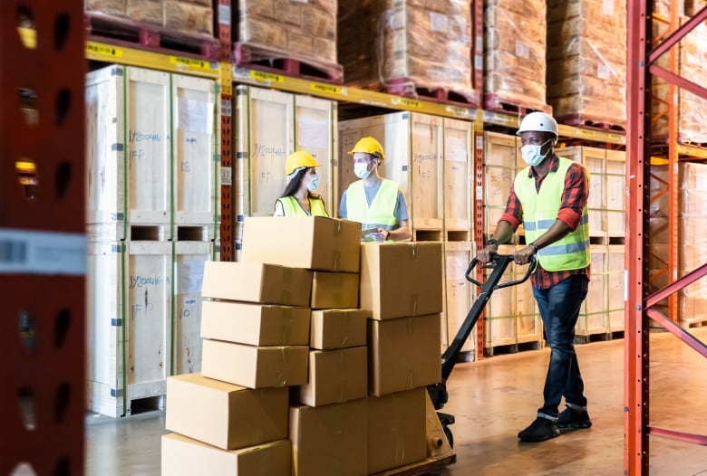 Group of workers wearing protective face masks working in factory warehouse. One is pushing a metal cart containing boxes.