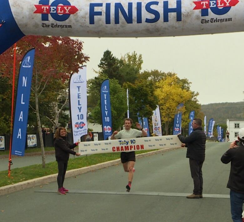 A man crosses the finish line. He is about to break the paper ribbon two people are holding up.