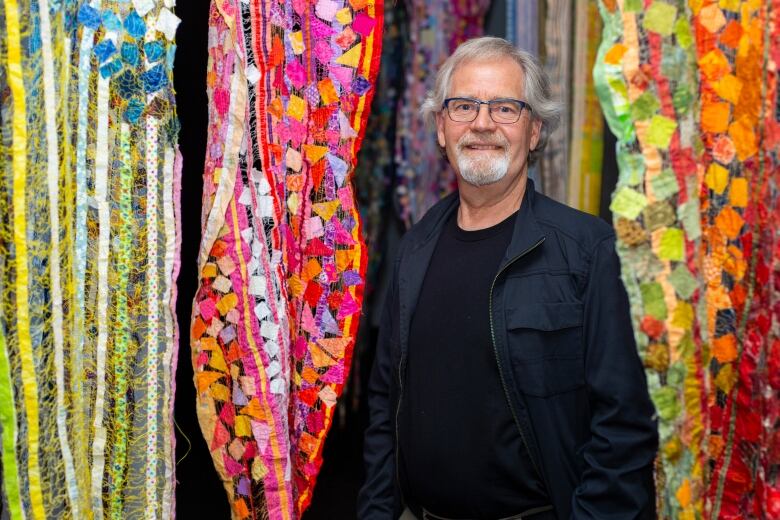 A man with white hair and a goatee stands surrounded by colourful fabric.