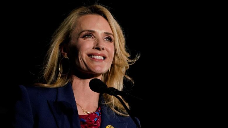 Close up of a smiling individual with long blond hair, wearing a red top and blue blazer.