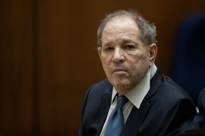 Close up of an unsmiling individual sitting at courtroom table wearing a blue suit and blue tie.