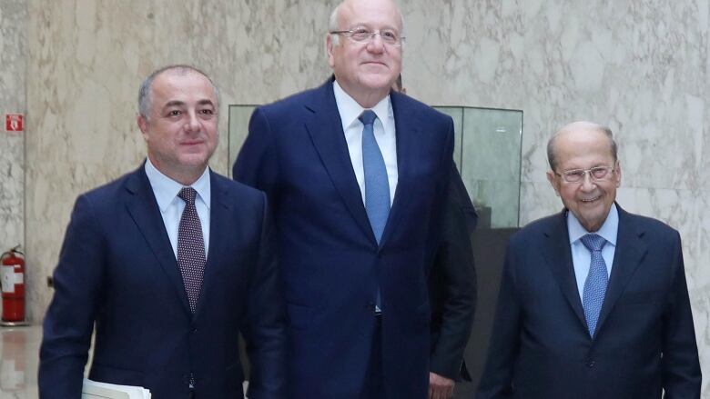 Three individuals, all grey-haired and balding, wearing blue suits, stand side by side. 