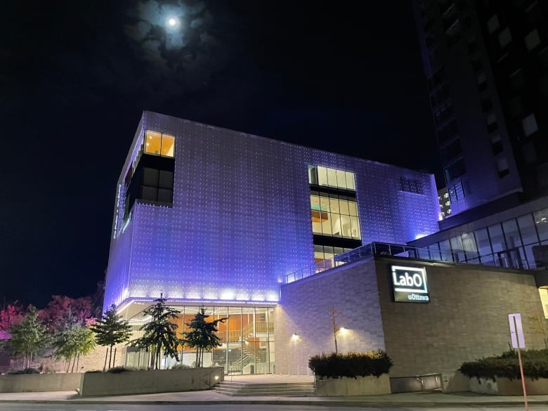 A square building bathed in purple at night. A nearly full moon is behind it.