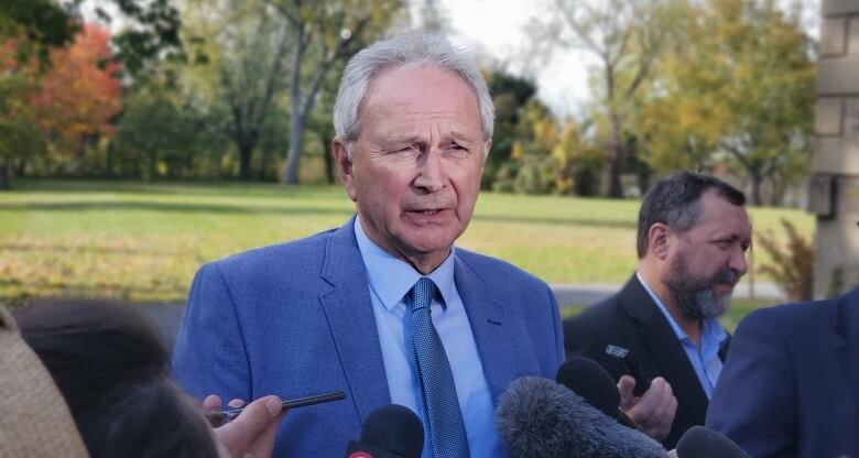 man in blue suit talks to a group of reporters outside.