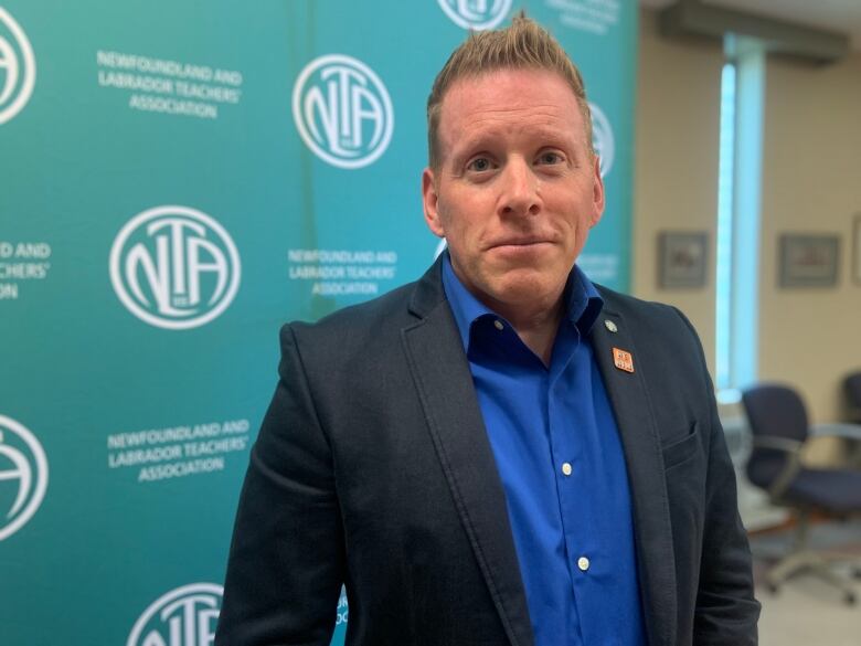 A medium close up of a person wearing a blue shirt and black jacket. The person is looking at the camera and standing in front of a green banner with the NLTA logo.