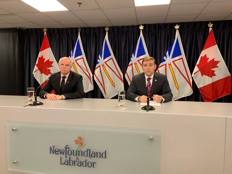 Two men wearing suits sit at a desk during a press conference.