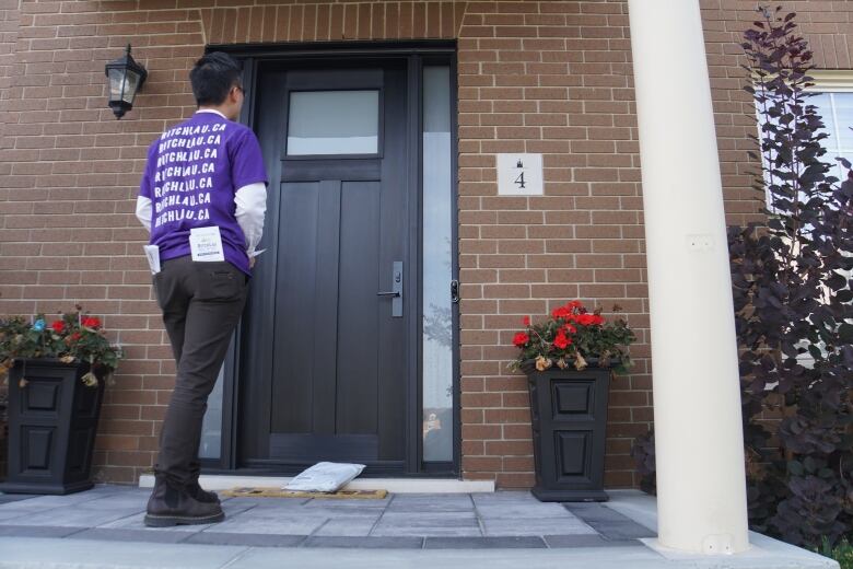 Markham council candidate Ritch Lau out door knocking, standing in a purple campaign shirt out front of a black door, waiting for its resident to answer.