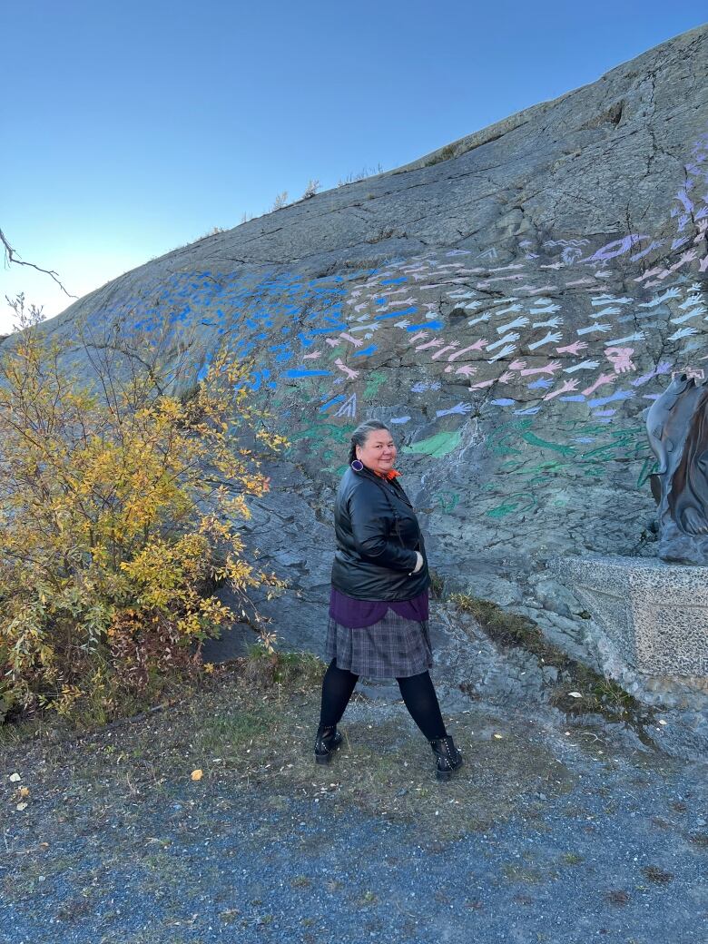 A portrait of a woman posing outdoors near a mural painted on a rocky cliff face.