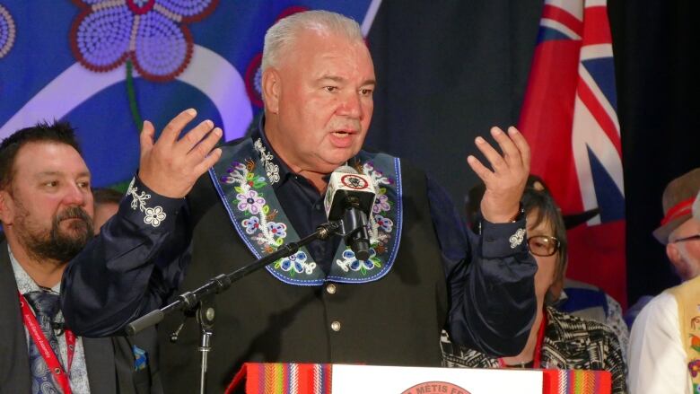 A white-haired man stands at a podium with his arms raised.