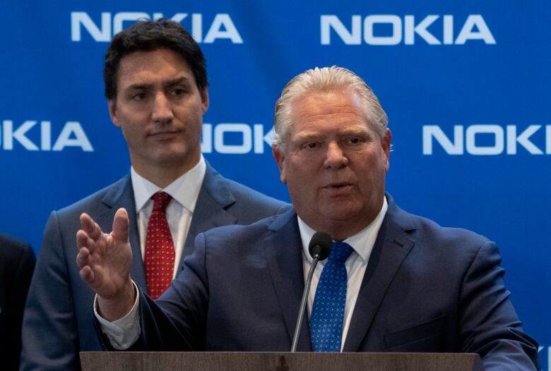 A politician speaks at a lectern in front of a blue corporate backdrop while another listens behind him.